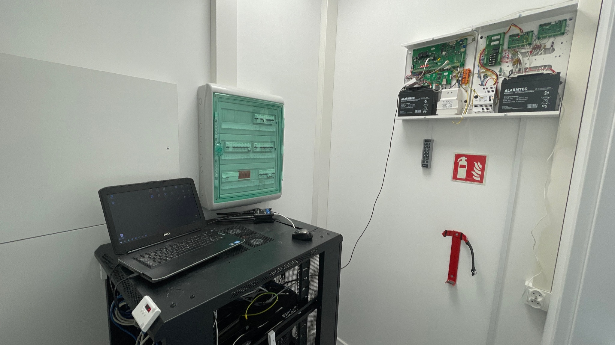 Czarny laptop Dell na metalowym regale obok zielonej skrzynki bezpieczników, na ścianie biała obudowa z elektroniką alarmową i akumulatorami Alarmtec, obok klawiatura numeryczna, znak ppoż i uchwyt...