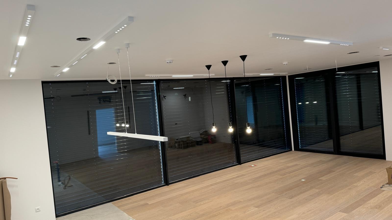 Nowoczesne biuro z drewnianą podłogą i dużymi oknami z żaluzjami, oświetlone listwami LED i wiszącymi lampami z żarówkami Edisona, widoczne otwory w suficie przed montażem opraw.