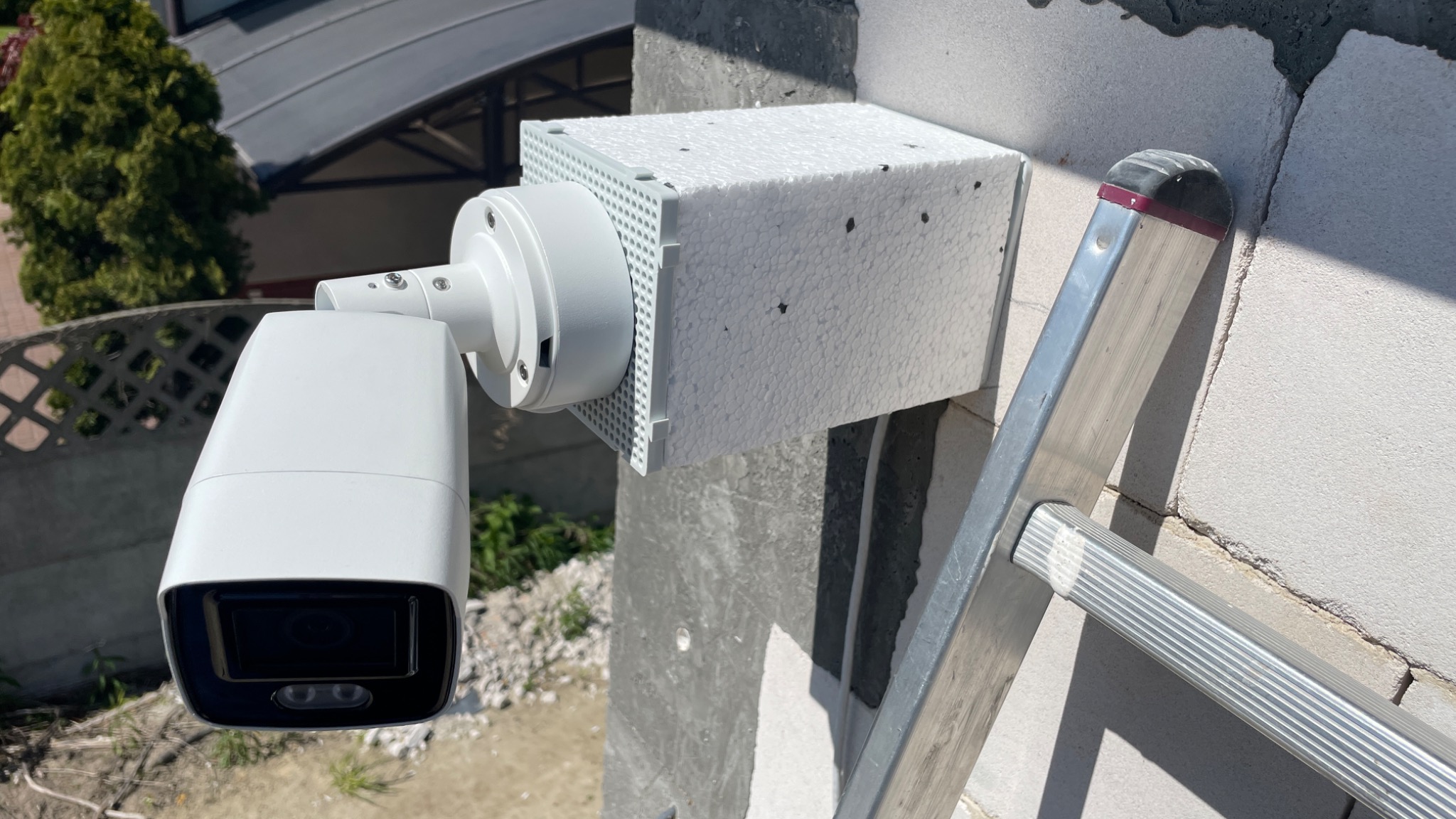 Biała kamera monitoringu zewnętrznego zamontowana na ścianie z bloczków betonowych, zabezpieczona styropianową obudową, widoczna drabina oparta o ścianę.