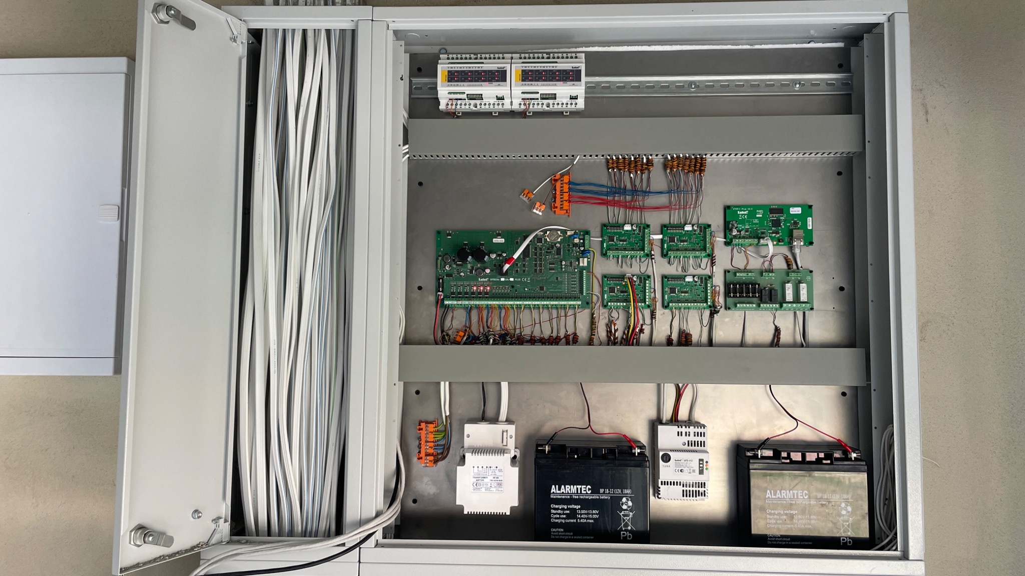 Otwarta szafa sterownicza z modułami alarmowymi, zasilaczami awaryjnymi i dużą wiązką kabli.