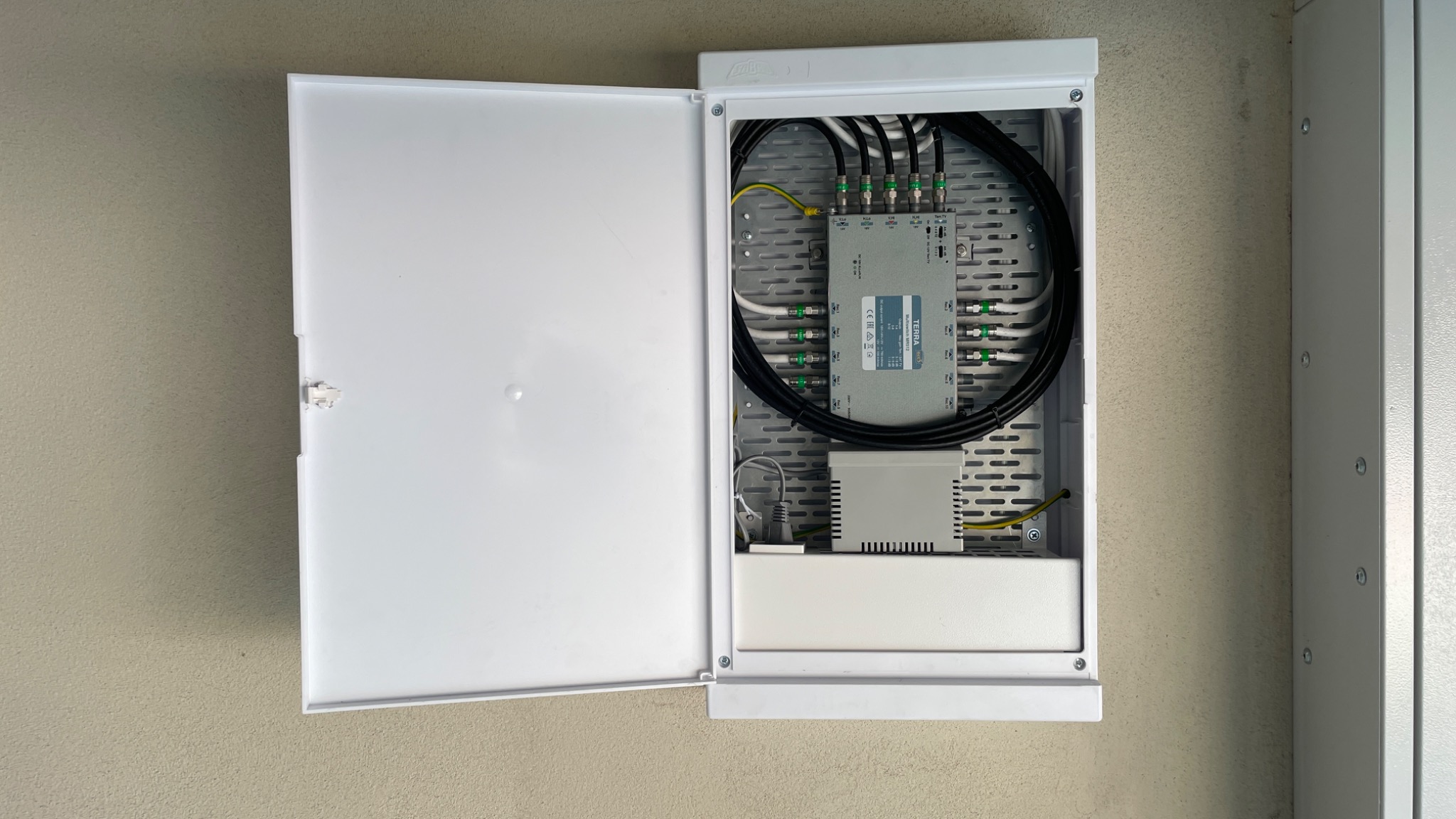 Otwarta, biała skrzynka z rozdzielnicą kablową na ścianie, widoczne kable koncentryczne i zasilające, oraz urządzenie Terra MTS4.