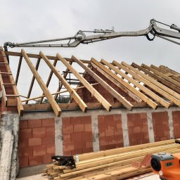 EKO BUD group - Wznoszenie ścian domu z czerwonej cegły ceramicznej, widoczne drewniane elementy konstrukcji dachu oraz ramię pompy do betonu i niwelator optyczny.