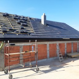 EKO BUD group - Budowa domu: widok na częściowo pokryty dach czarną dachówką, komin z pustaków, ściany z czerwonej cegły, rusztowanie na kółkach z narzędziami i butelką wody, drabina oparta o ścianę, deski...