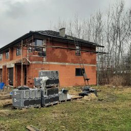 EKO BUD group - Niedokończony dwukondygnacyjny dom murowany z czerwonej cegły, z widocznymi rusztowaniami, paletami z materiałami budowlanymi marki BMI Braas oraz metalową drabiną opartą o ścianę, na tle...