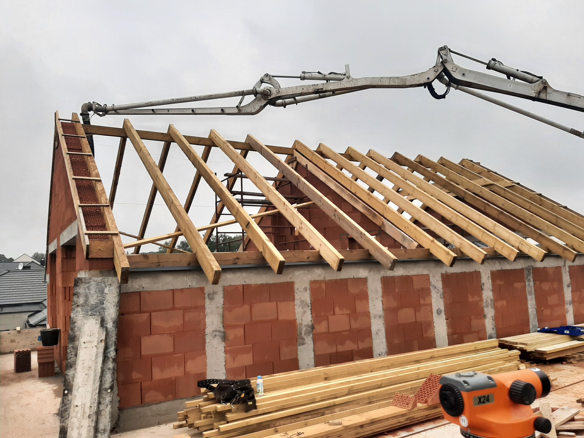 Wznoszenie ścian domu z czerwonej cegły ceramicznej, widoczne drewniane elementy konstrukcji dachu oraz ramię pompy do betonu i niwelator optyczny.