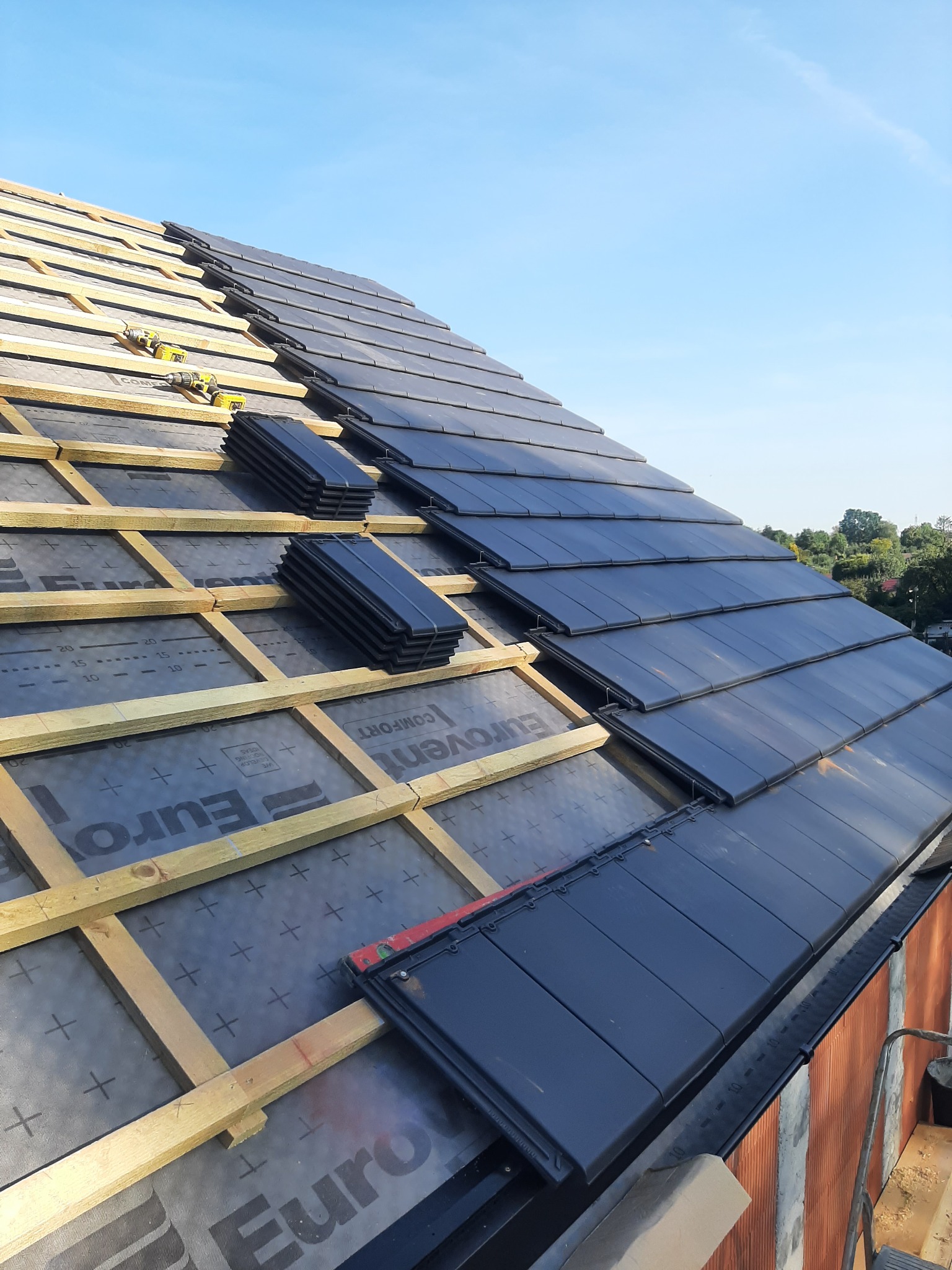 Układanie czarnej dachówki na nowym dachu z widoczną membraną Eurovent, łatami i kontrłatami oraz dwoma wkrętarkami akumulatorowymi Ryobi na połaci.