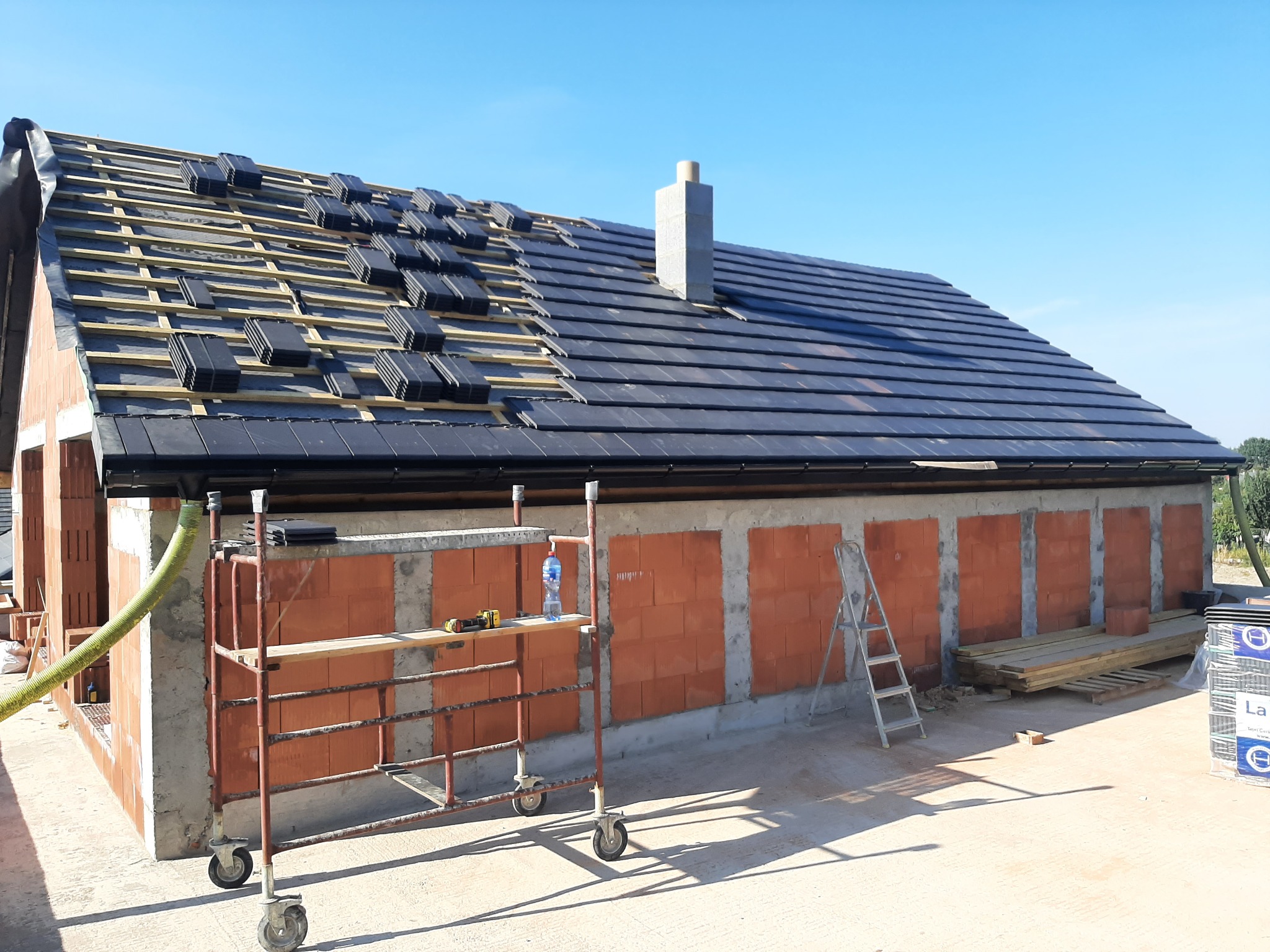 Budowa domu: widok na częściowo pokryty dach czarną dachówką, komin z pustaków, ściany z czerwonej cegły, rusztowanie na kółkach z narzędziami i butelką wody, drabina oparta o ścianę, deski...