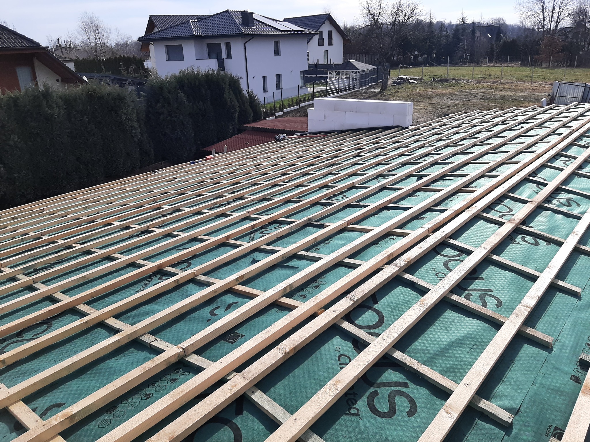 Konstrukcja drewniana dachu pokryta zieloną membraną hydroizolacyjną, widoczne domy jednorodzinne w tle.