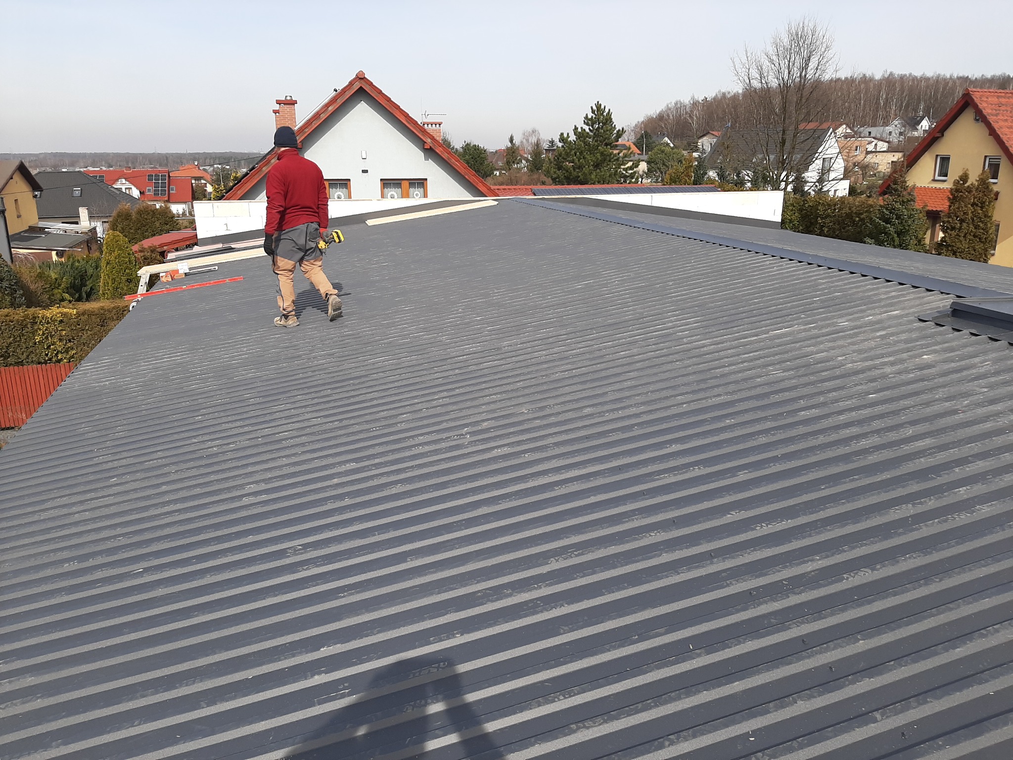 Monter w czerwonej bluzie idzie po nowo położonym, szarym dachu z blachy trapezowej, trzymając w ręce wkrętarkę; w tle zabudowa jednorodzinna i bezlistne drzewa.