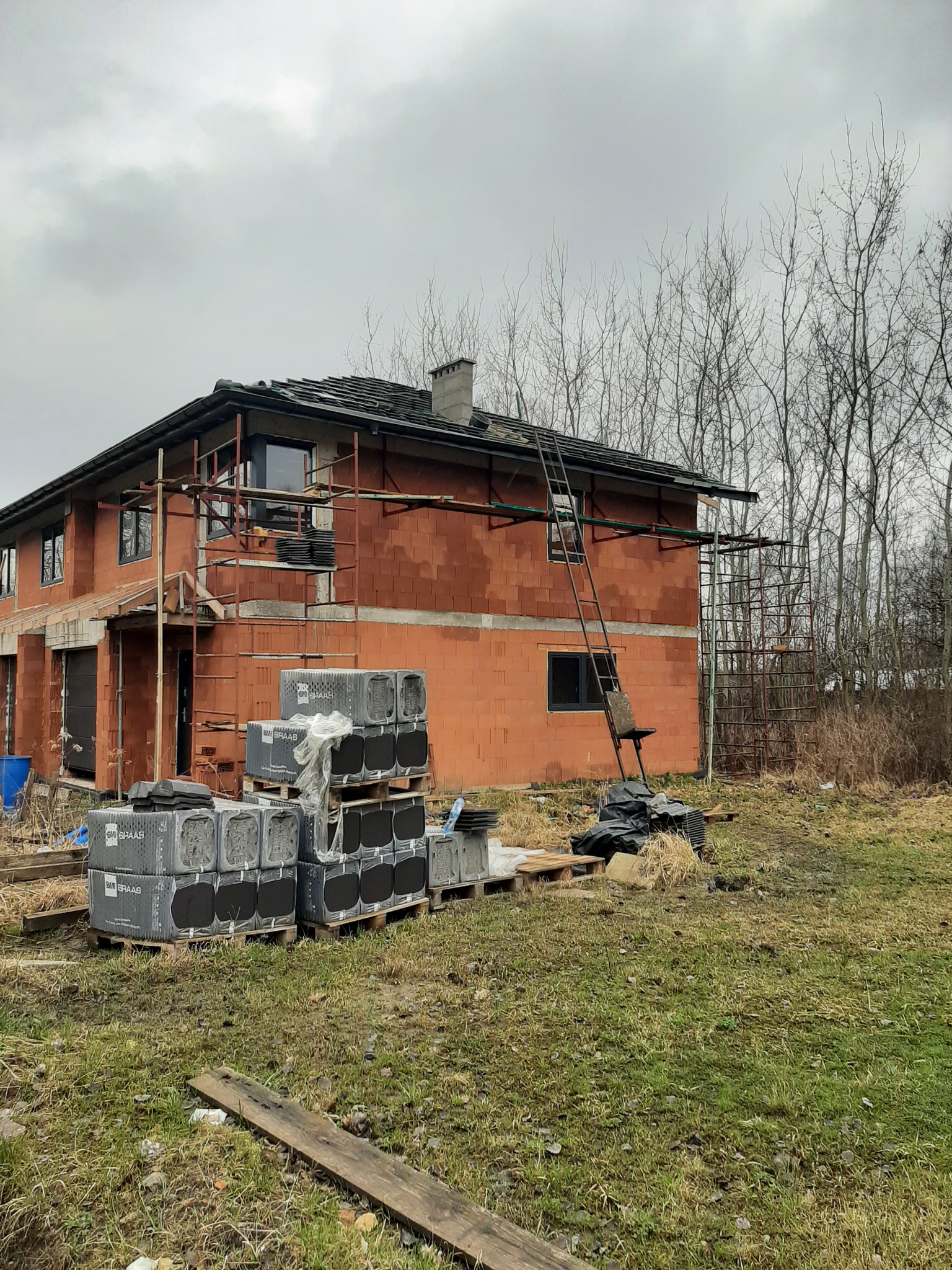 Niedokończony dwukondygnacyjny dom murowany z czerwonej cegły, z widocznymi rusztowaniami, paletami z materiałami budowlanymi marki BMI Braas oraz metalową drabiną opartą o ścianę, na tle...