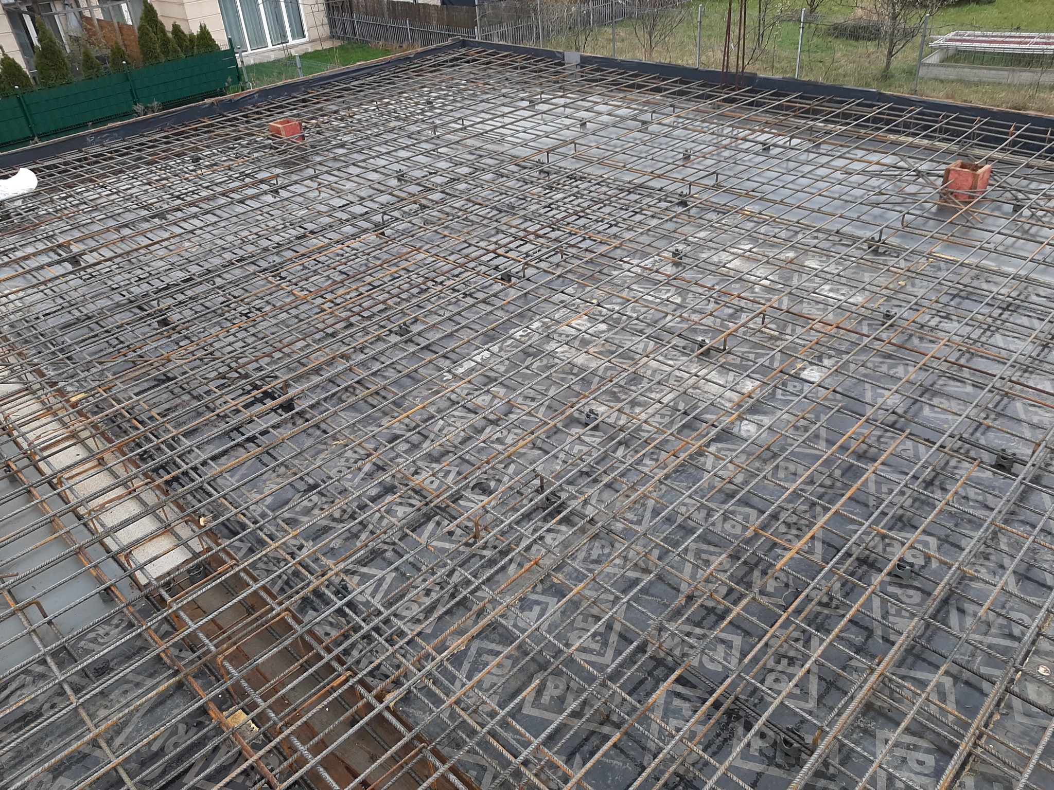 Siatka zbrojeniowa na czarnej folii z widocznymi podkładkami dystansowymi, przygotowana do wylania betonu, z cegłą na krawędzi i zielenią w tle.