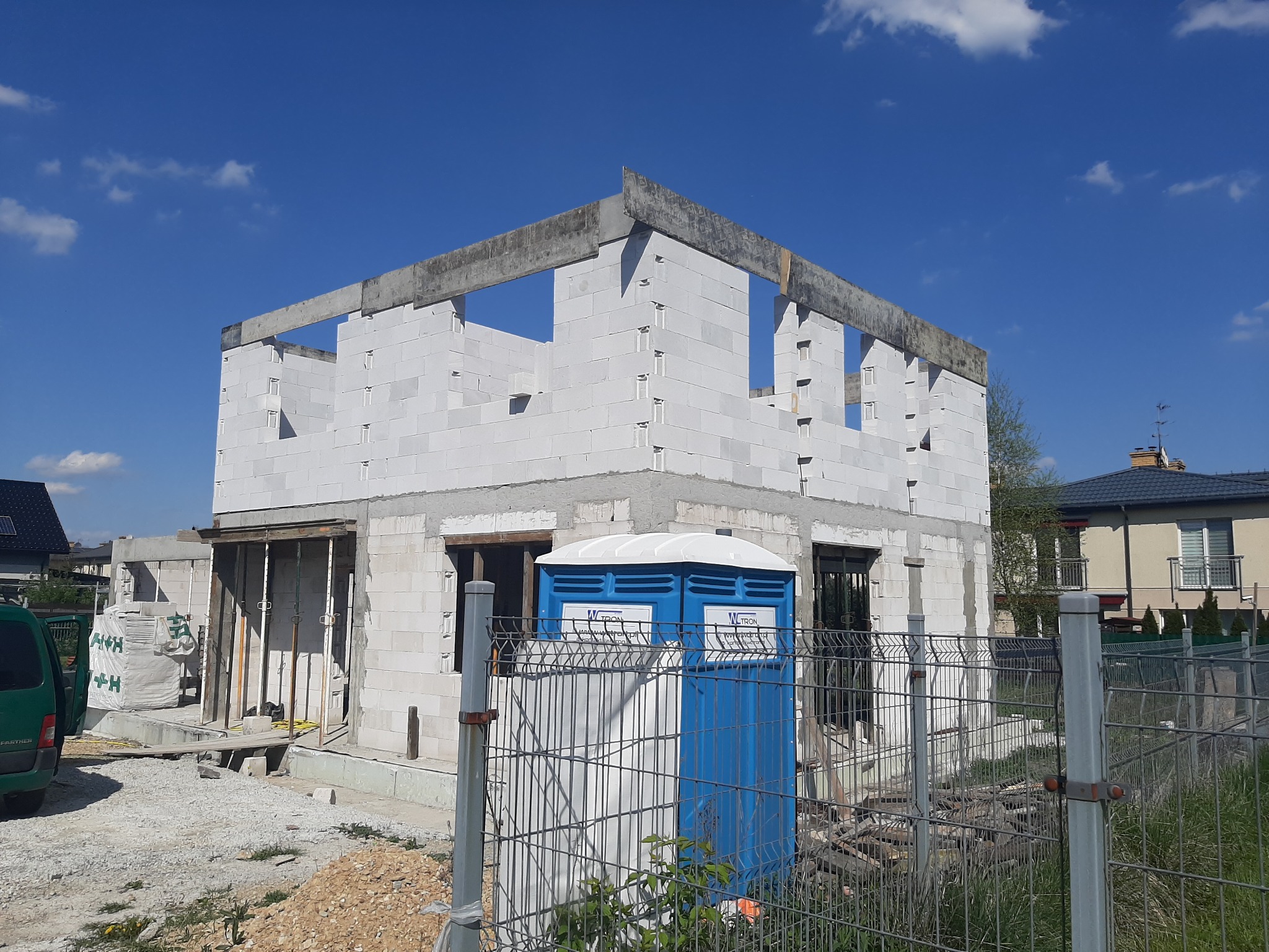 Surowy stan domu z białych bloczków, widoczne otwory okienne, betonowy wieniec, niebieska toaleta przenośna przed budynkiem, ogrodzenie z siatki, błękitne niebo.