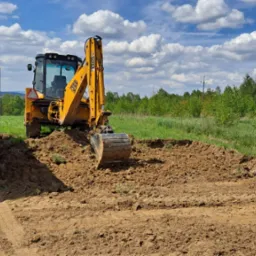 Żółta koparko-ładowarka JCB podczas prac ziemnych przy wykopie, obok stoi ciężarówka z przyczepą, w tle zielone drzewa i błękitne niebo z chmurami.
