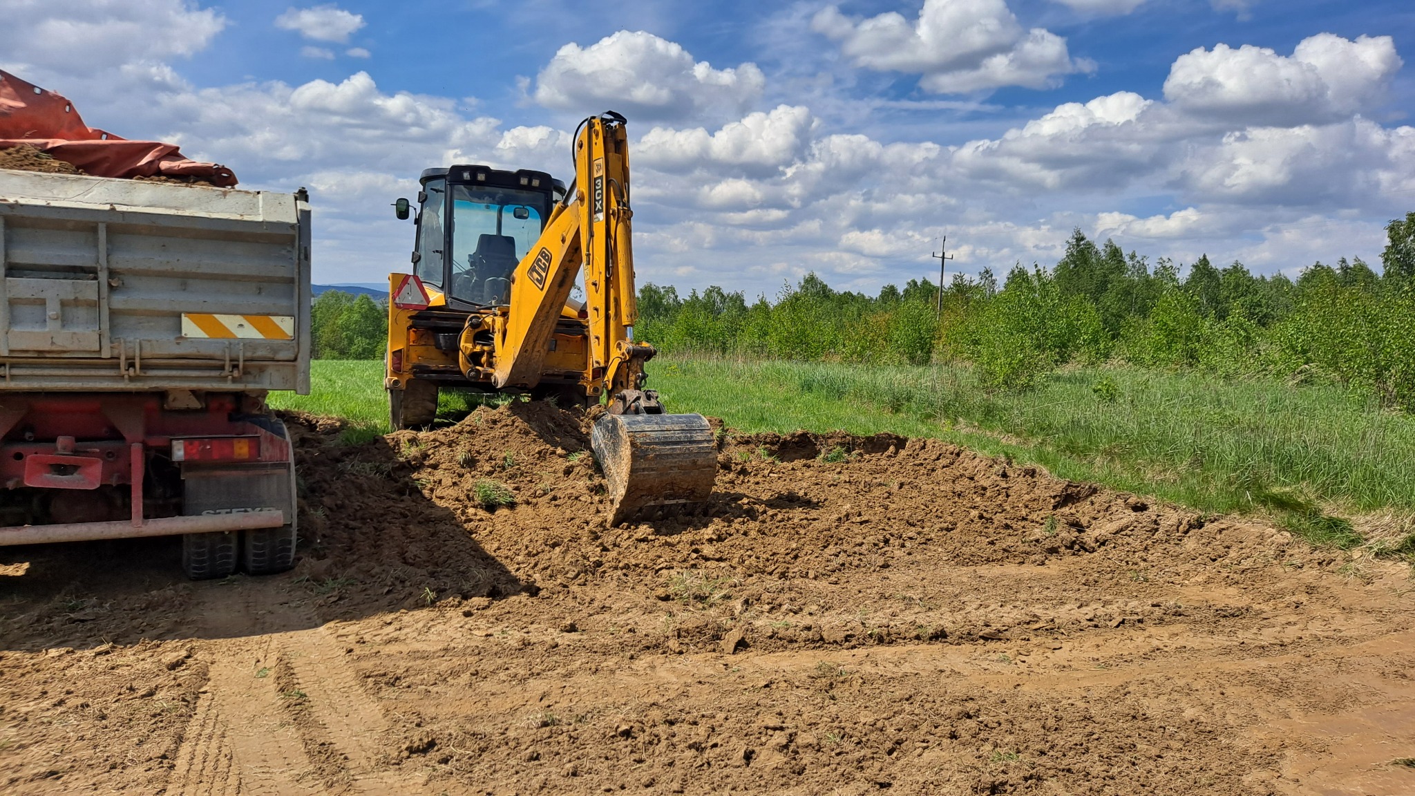 Żółta koparko-ładowarka JCB podczas prac ziemnych przy wykopie, obok stoi ciężarówka z przyczepą, w tle zielone drzewa i błękitne niebo z chmurami.