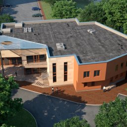PRIMALINEPROJEKT Sp. z o. o. - Widok z góry na nowoczesny budynek usługowy z płaskim dachem, szklanym zadaszeniem nad wejściem i elewacją w odcieniach beżu i pomarańczy, otoczony zielenią i parkingiem z kilkoma samochodami.