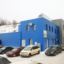 PRIMALINEPROJEKT Sp. z o. o. - Niebiesko-szary budynek usługowy z blachy falistej z rzędem zaparkowanych samochodów na śniegu, widoczne okna łukowe i tablica reklamowa.