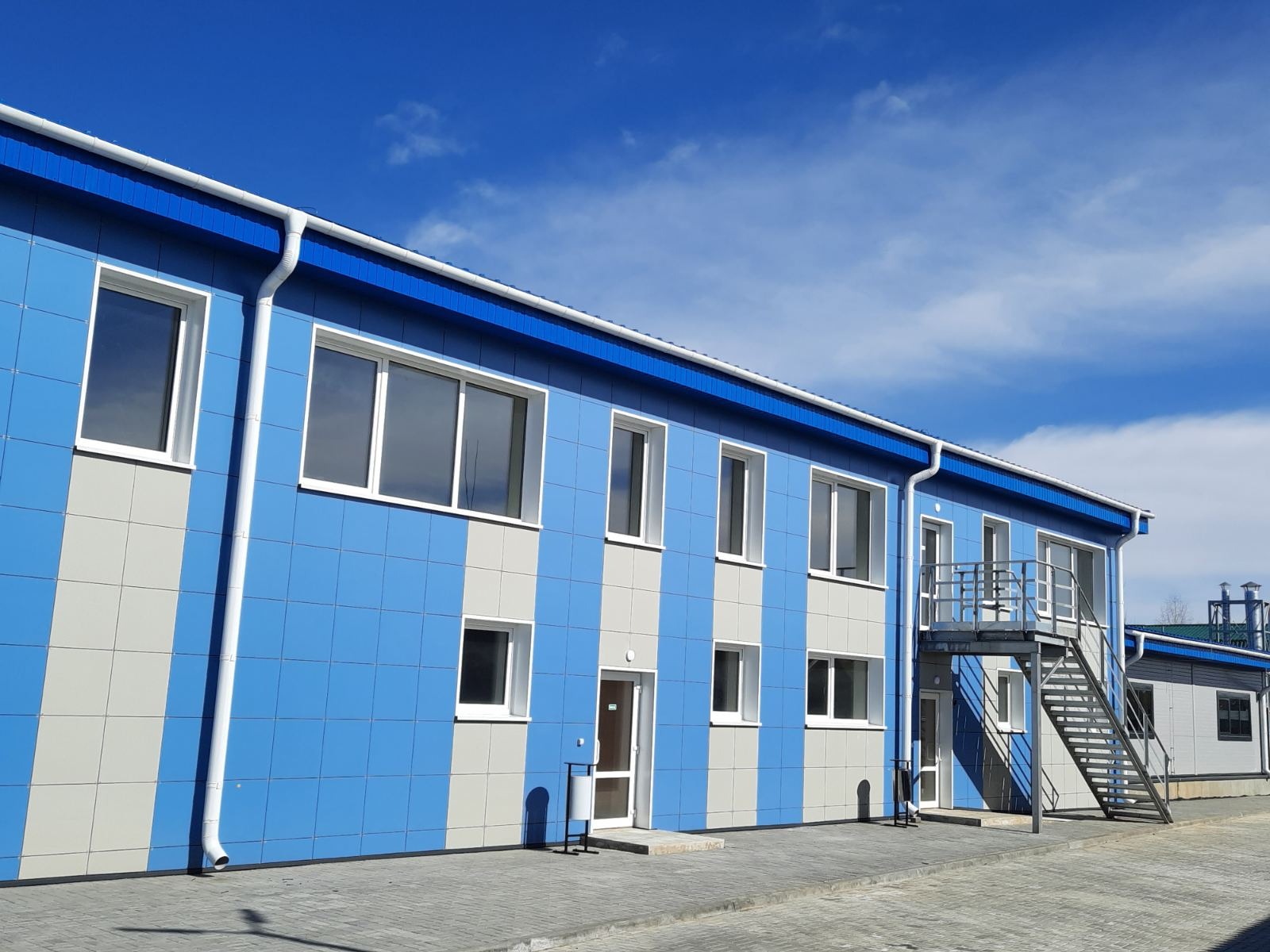 Nowoczesny budynek usługowy z niebiesko-szarą elewacją w pionowe pasy, białe okna i zewnętrzne schody ewakuacyjne, na tle błękitnego nieba.