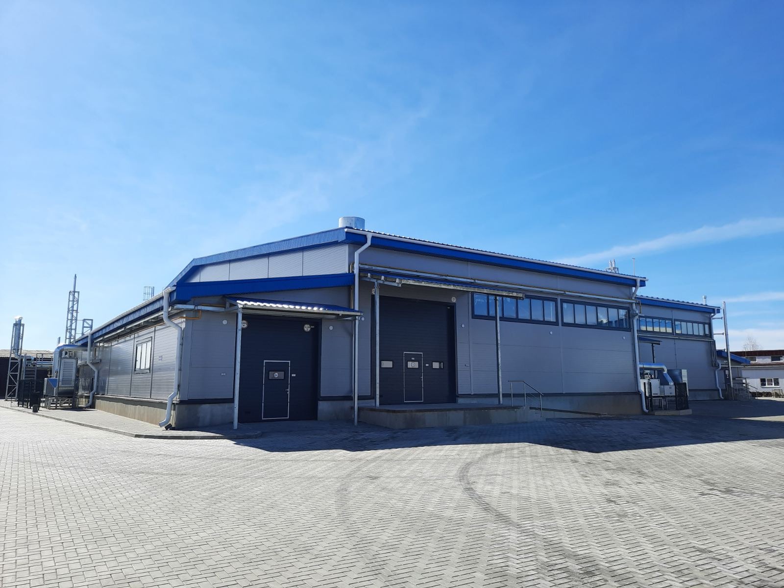 Nowoczesna hala przemysłowa z szarymi ścianami, niebieskim dachem i dwiema bramami wjazdowymi, widok z zewnątrz na tle błękitnego nieba.