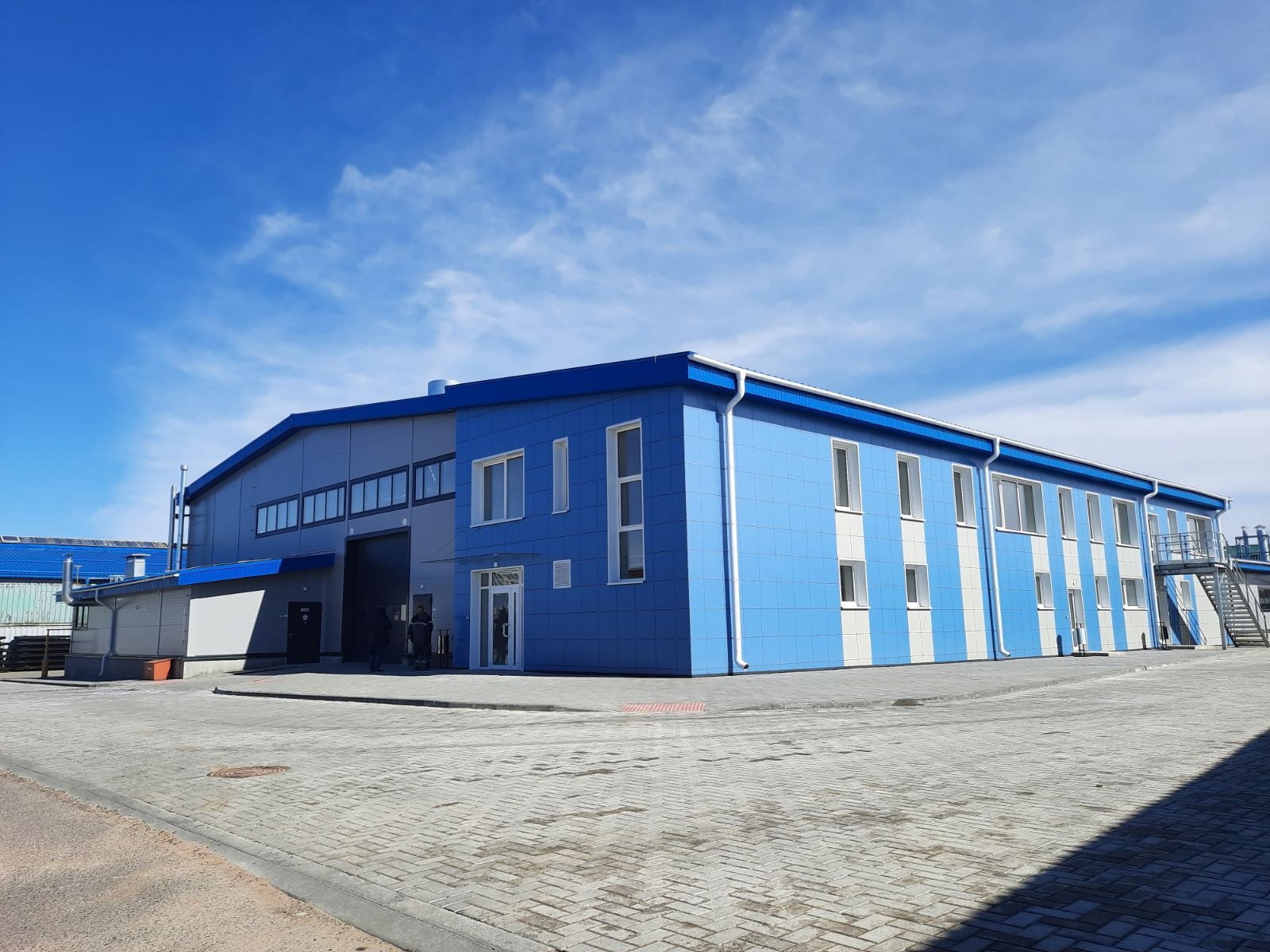 Nowoczesna hala produkcyjna w odcieniach błękitu i szarości, z białymi akcentami, widziana z zewnątrz w słoneczny dzień. Na pierwszym planie brukowa nawierzchnia.