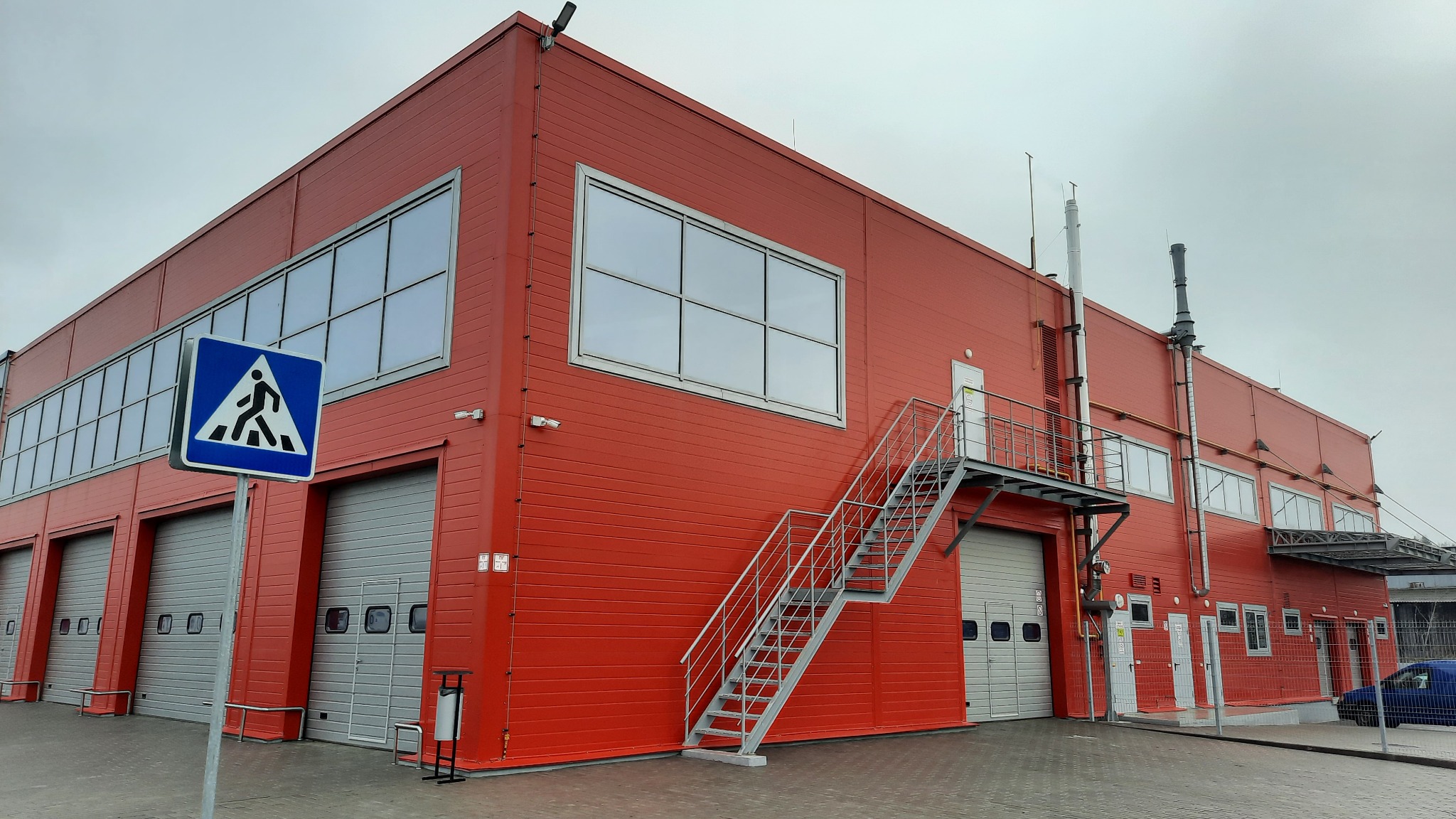 Czerwony budynek przemysłowy z bramami garażowymi, oknami i zewnętrznymi schodami stalowymi, widziany z poziomu chodnika, z niebieskim znakiem przejścia dla pieszych w pierwszym planie.