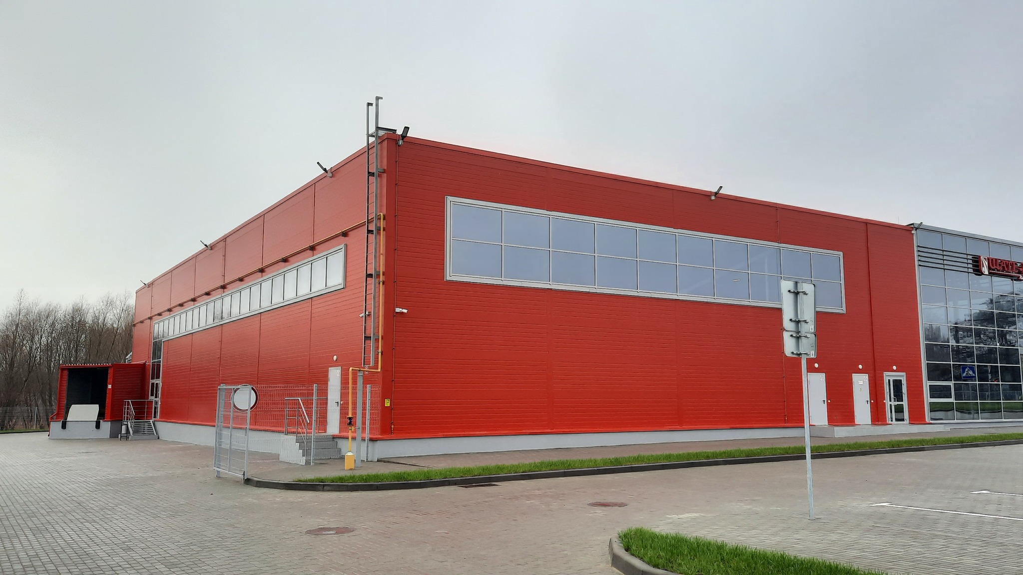Czerwona hala magazynowa z panelowymi ścianami i rzędem okien, widoczna rampa załadunkowa i boczna drabina.