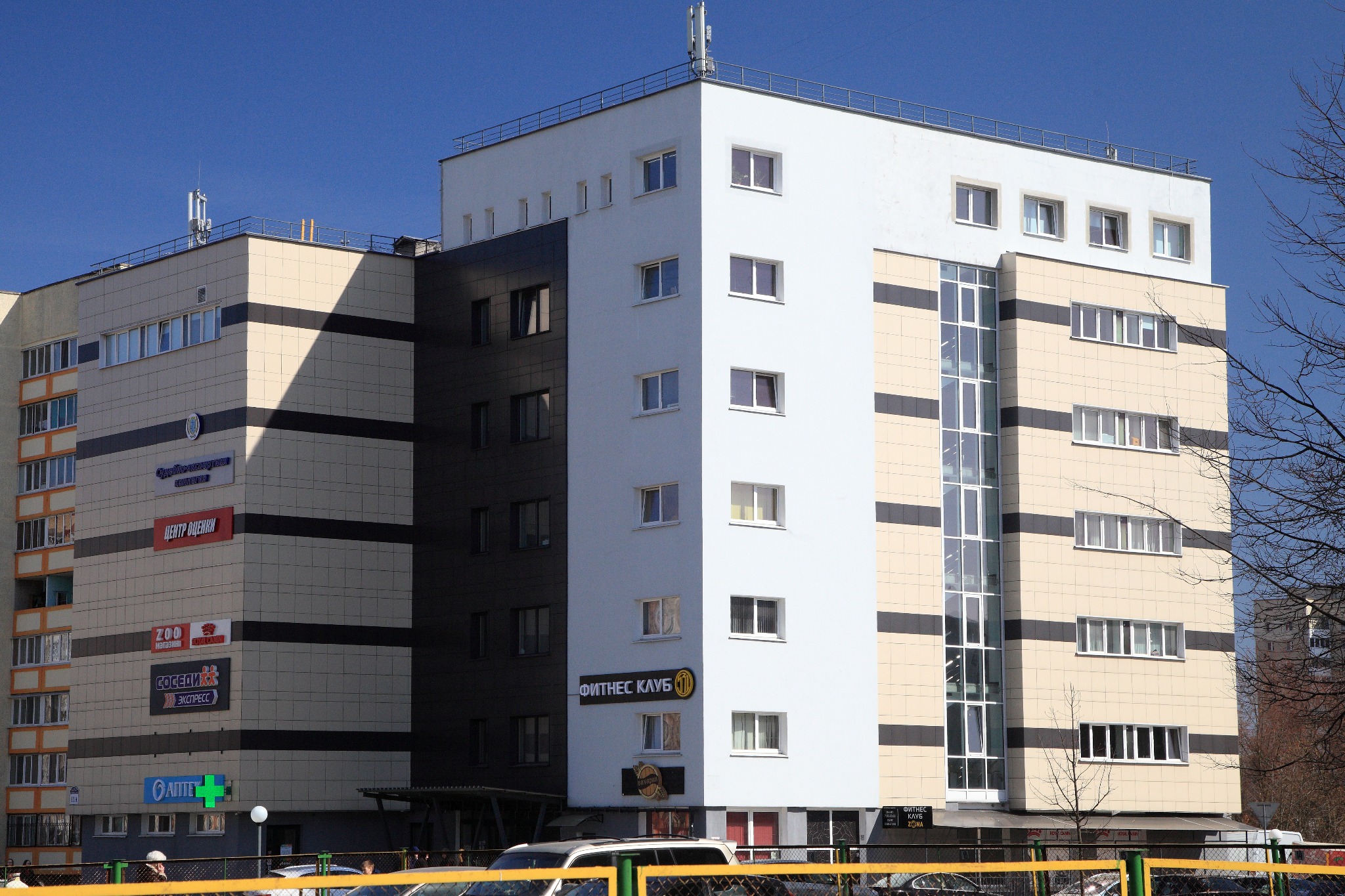 Wielopiętrowy budynek usługowy z panelami w kolorach beżowym, białym i czarnym, z oknami i reklamami firm na fasadzie, widziany z poziomu ulicy w słoneczny dzień.