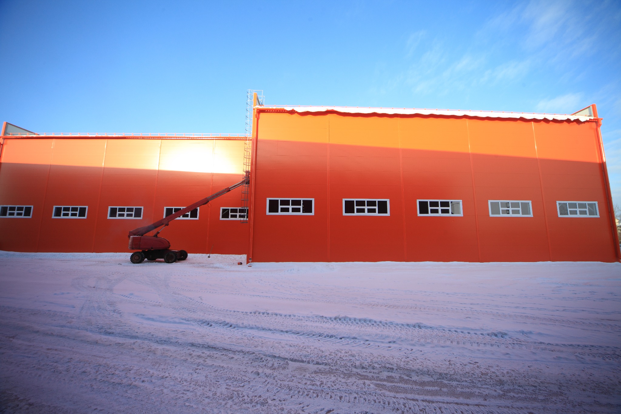 Pomarańczowa hala przemysłowa z rzędem okien i podnośnikiem koszowym na tle zimowego krajobrazu.