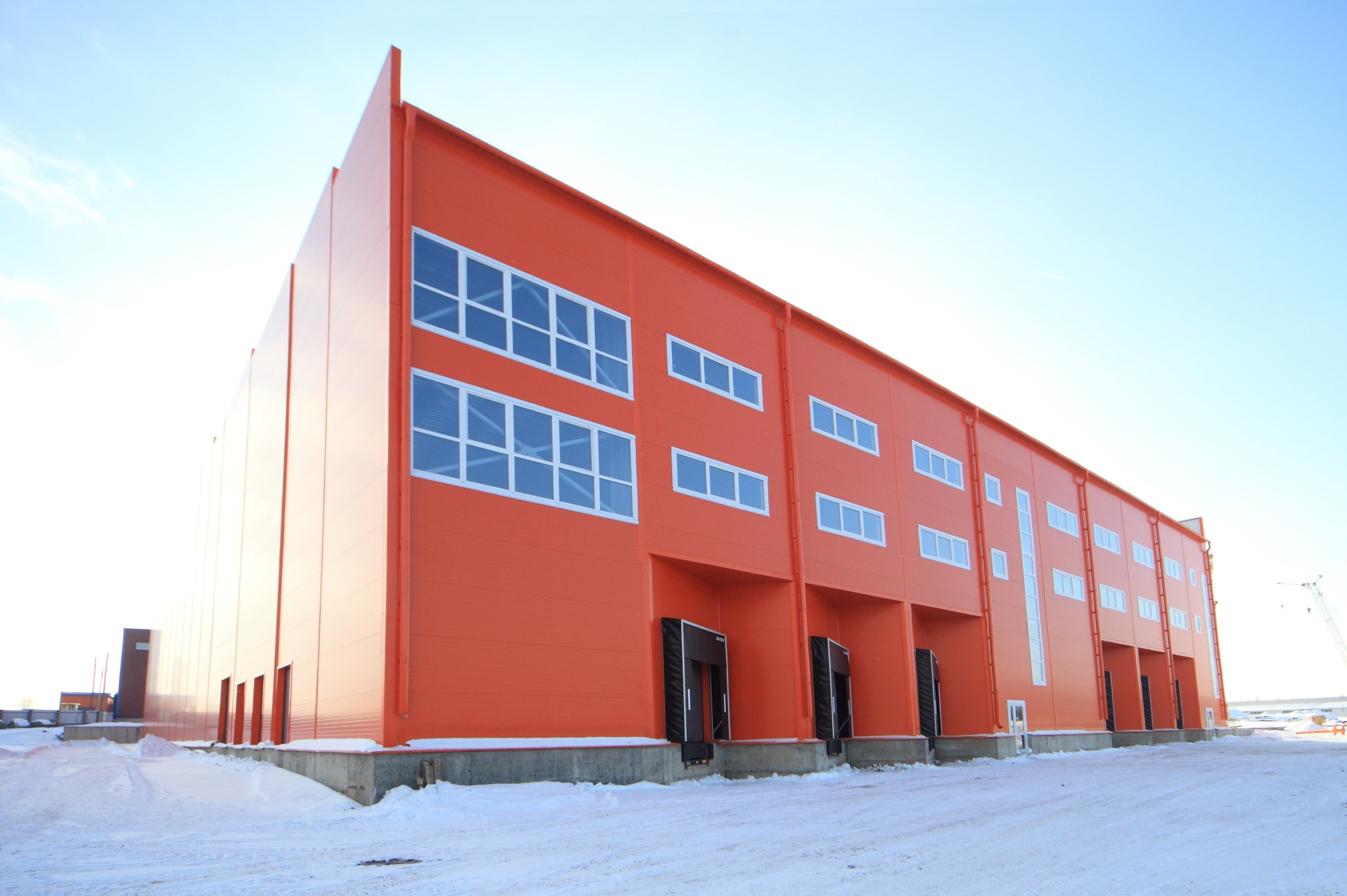 Pomarańczowa hala przemysłowa z białymi oknami i czarnymi bramami wjazdowymi, otoczona śnieżnym terenem w słoneczny dzień.