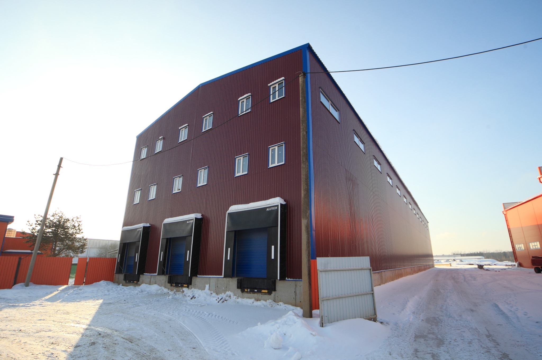 Zimowy krajobraz z widokiem na dużą, brązową halę magazynową z niebieskimi akcentami, wyposażoną w trzy bramy dokowe Alutech, oświetloną przez jasne, zimowe słońce. Ślady opon na śniegu prowadzą...