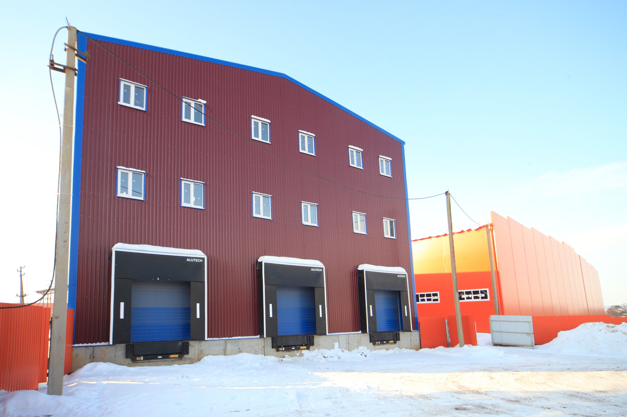 Gotowa hala magazynowa z bordową elewacją z blachy trapezowej, niebieskimi obramowaniami i trzema bramami segmentowymi Alutech, otoczona śniegiem w słoneczny dzień.