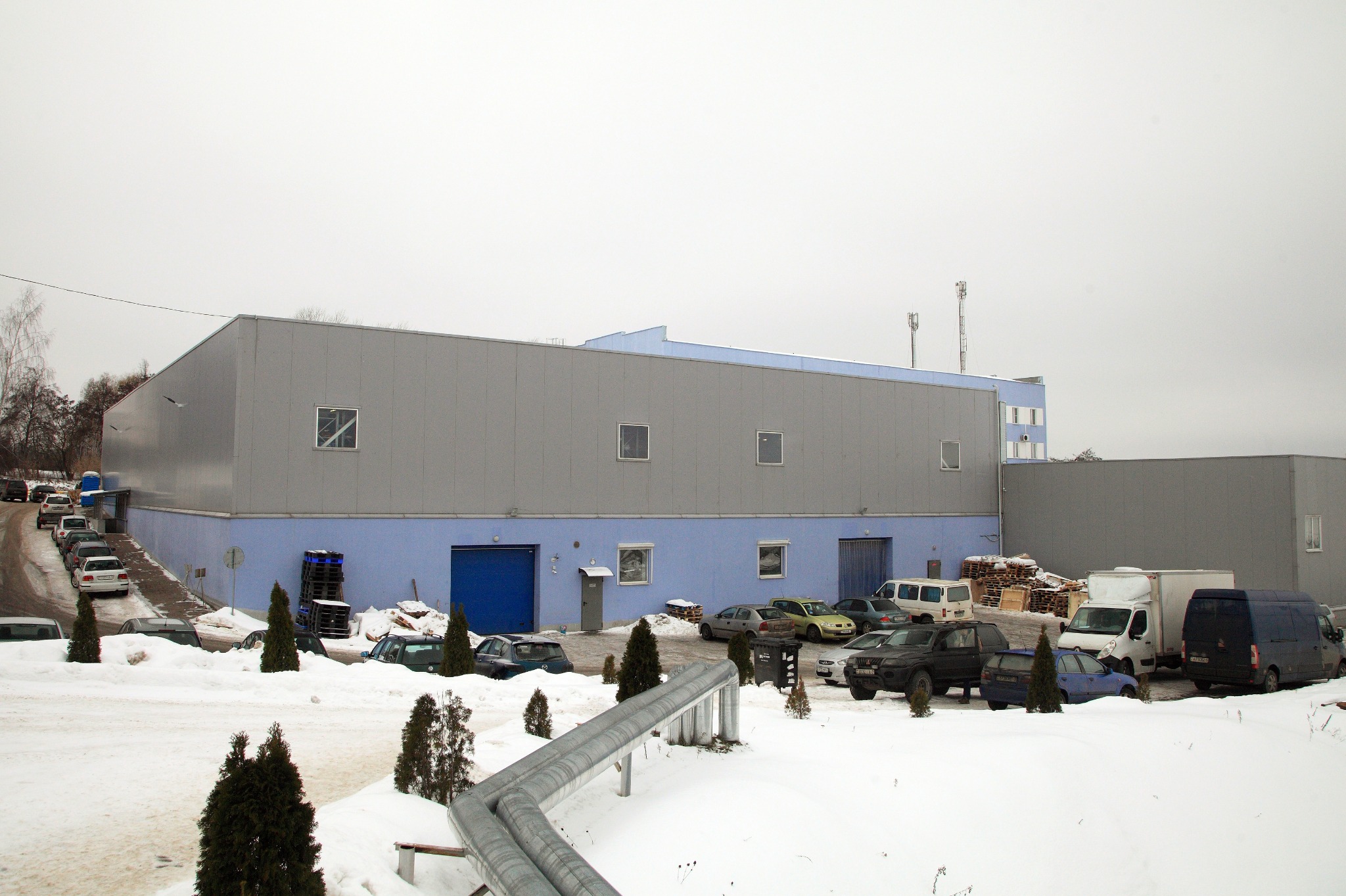 Zewnętrzny widok hali produkcyjnej z szarej blachy trapezowej i niebieskiego betonu, otoczonej śniegiem, z zaparkowanymi samochodami i widocznymi rurami ciepłowniczymi na pierwszym planie.