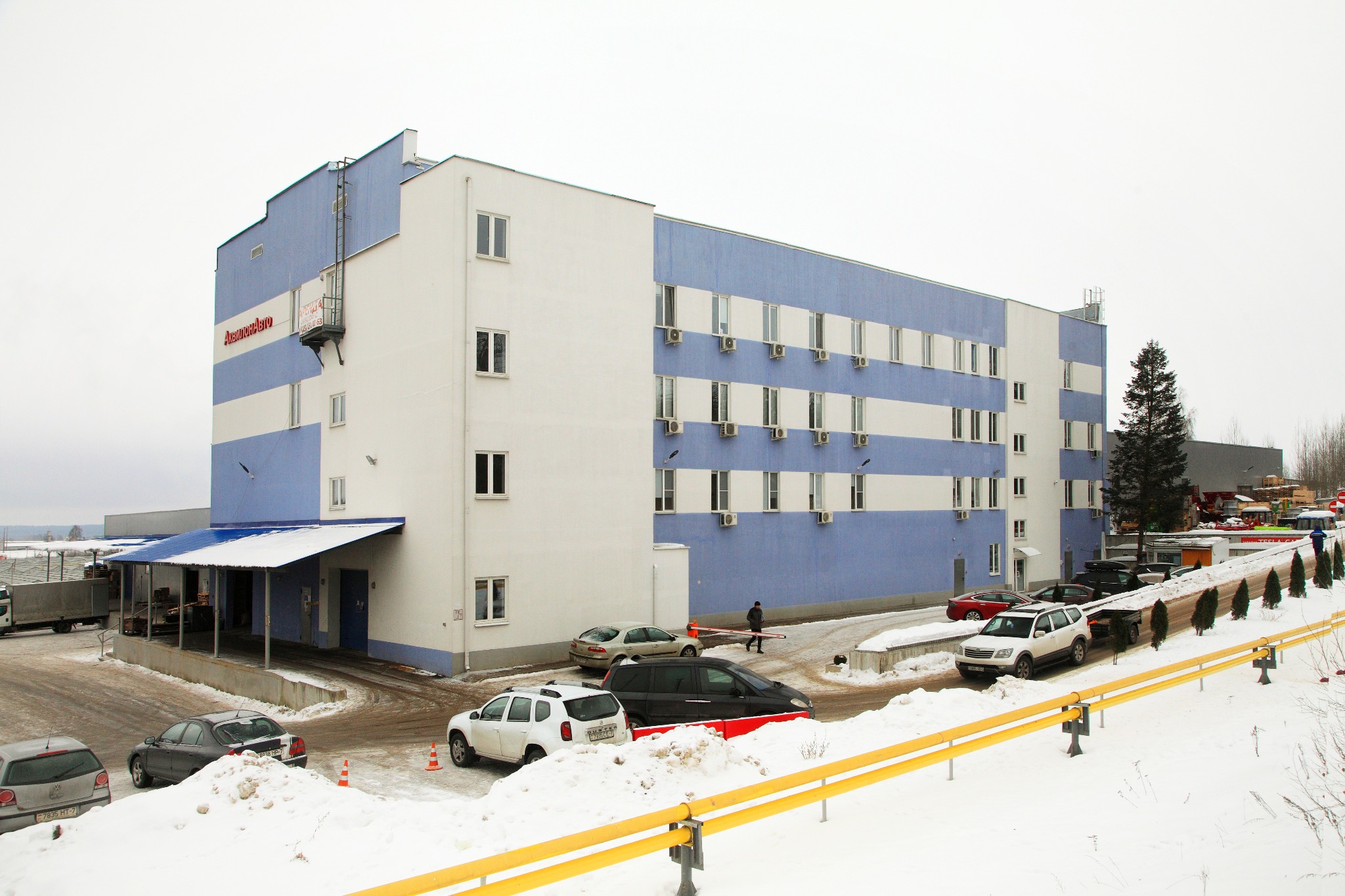 Zewnętrzny widok na dwukolorowy budynek usługowy z płaskim dachem, z zaparkowanymi samochodami i zaśnieżonym otoczeniem w zimowy dzień.