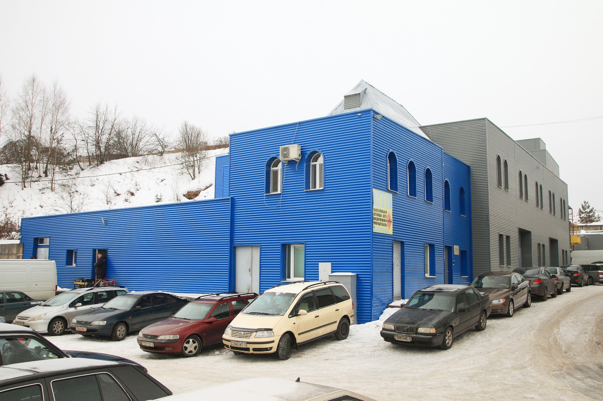 Niebiesko-szary budynek usługowy z blachy falistej z rzędem zaparkowanych samochodów na śniegu, widoczne okna łukowe i tablica reklamowa.