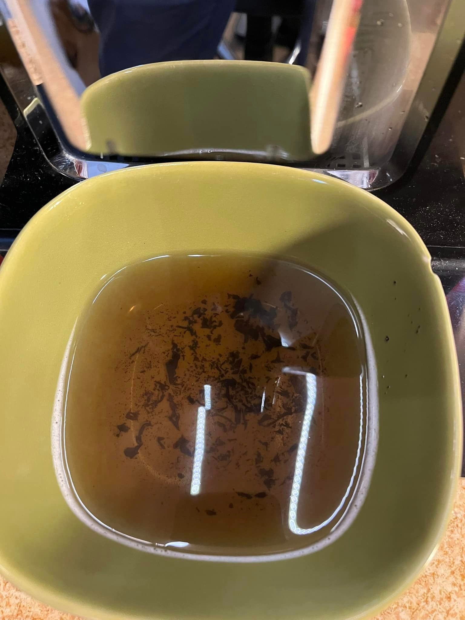 Żółta miseczka z brązowym płynem i fusami, odbicie ekspresu do kawy w powierzchni płynu.