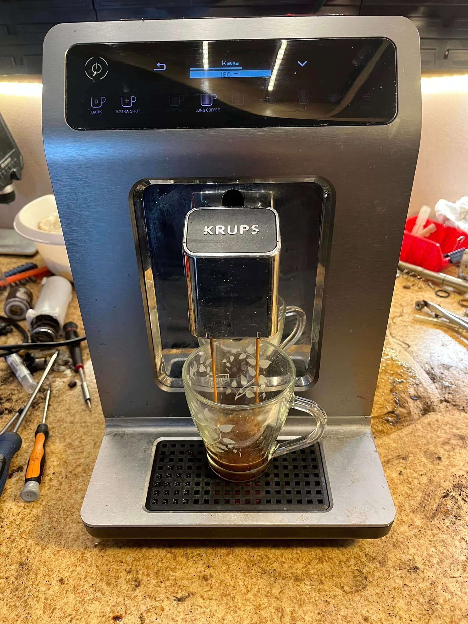 Szaro-czarny ekspres Krups w trakcie parzenia kawy do szklanki z delikatnym wzorem, widoczne narzędzia i części, sugerujące serwis lub naprawę w tle.