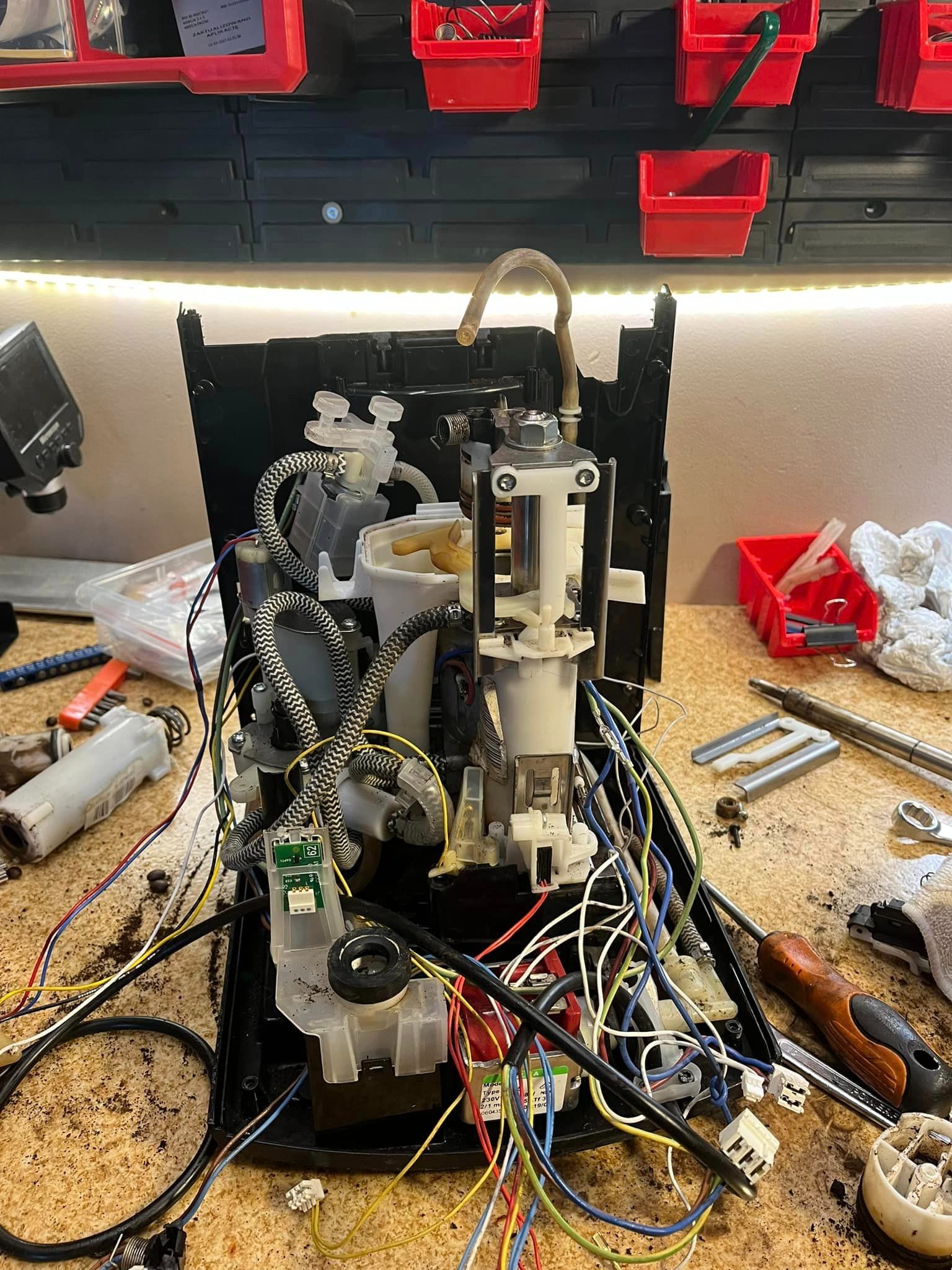 Rozłożony ekspres do kawy na warsztatowym stole, widoczne elementy wewnętrzne, kable, narzędzia i pojemniki z częściami.