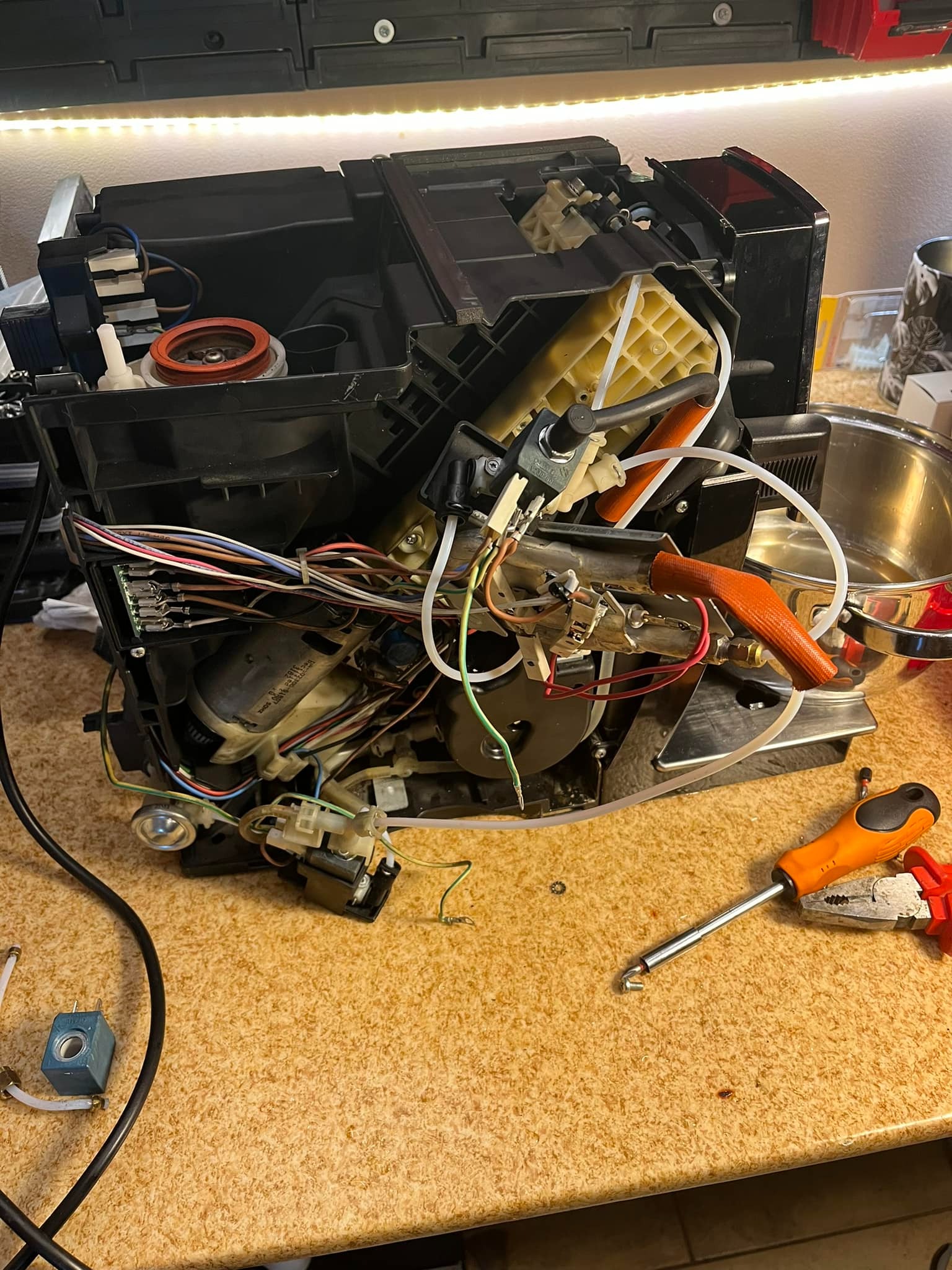 Rozłożony ekspres do kawy na stole podczas naprawy, widoczne elementy wewnętrzne, przewody, narzędzia takie jak śrubokręt i kombinerki.