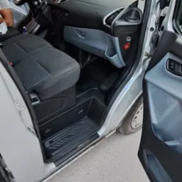 Wnętrze srebrnego samochodu dostawczego z otwartymi drzwiami, widoczne czarne fotele z szarym wzorem, deska rozdzielcza i elementy wyposażenia.