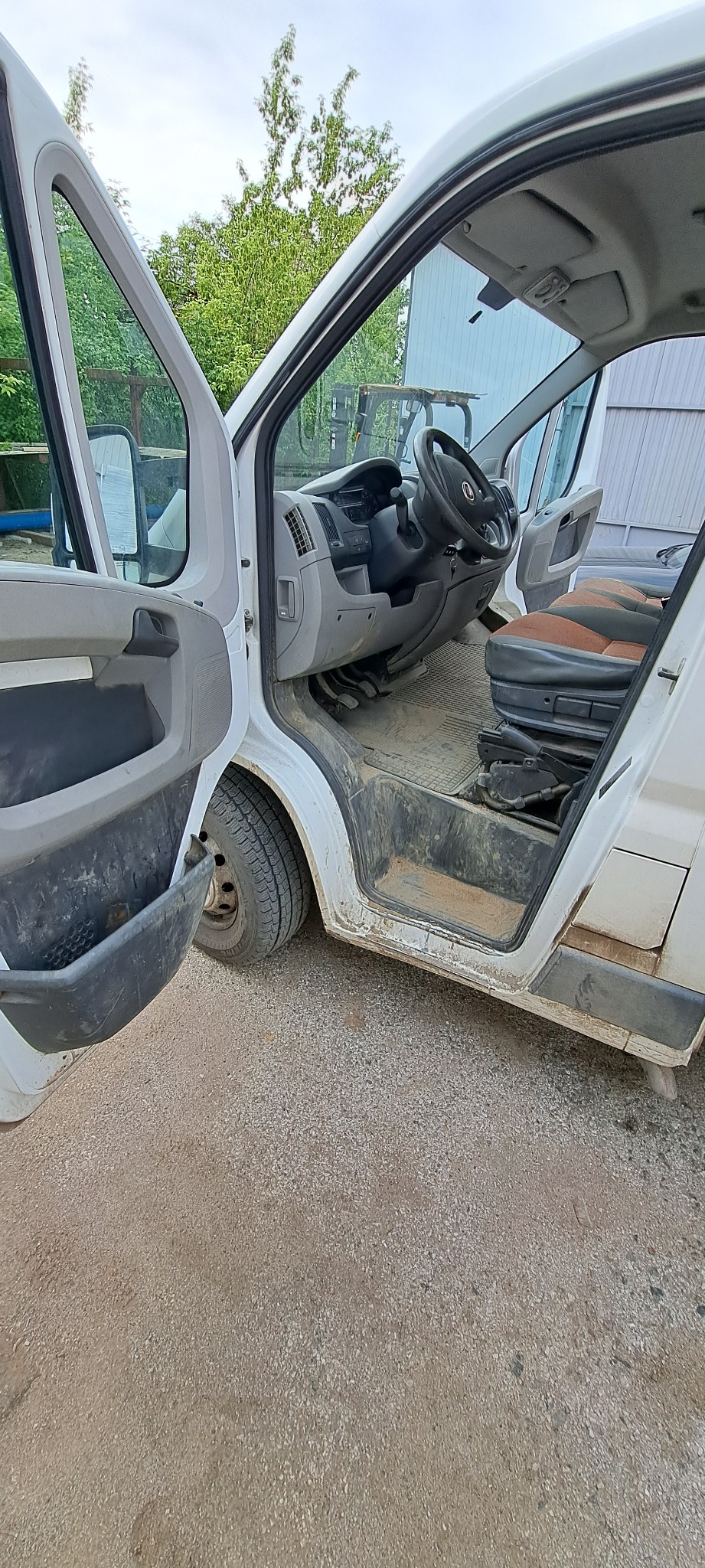 Wnętrze brudnego, białego busa z otwartymi drzwiami, widoczne z zewnątrz na żwirowym podłożu, z zanieczyszczoną tapicerką foteli i zabrudzeniami na progach.