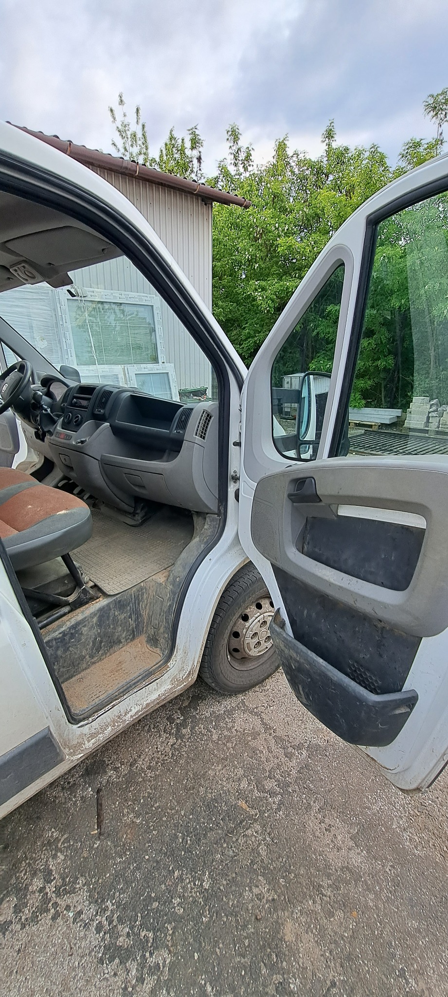 Brudny, biały samochód dostawczy z otwartymi drzwiami kierowcy, wnętrze zakurzone i zabrudzone, na zewnątrz betonowy plac i budynek gospodarczy w tle.