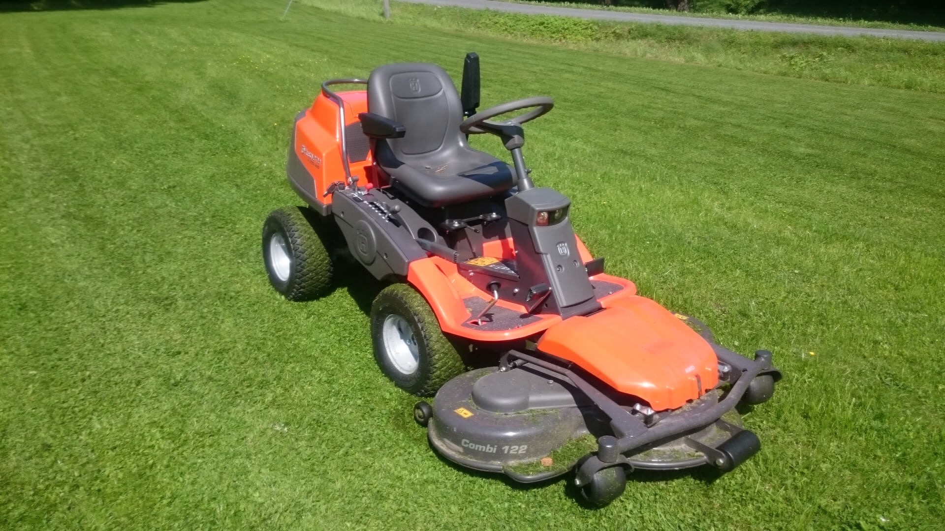 Pomarańczowo-szara kosiarka samojezdna Husqvarna na idealnie przystrzyżonym, zielonym trawniku w słoneczny dzień.