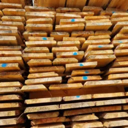 Stos równo ułożonych desek z widocznymi śladami cięcia i niebieskimi oznaczeniami na niektórych z nich, oświetlonych naturalnym światłem słonecznym.