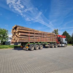 ADI-TRANS ADRIAN ZADKA - Ciężarówka z naczepą załadowana okrąglakami drewna na placu, widoczne zabezpieczenia ładunku, błękitne niebo w tle.