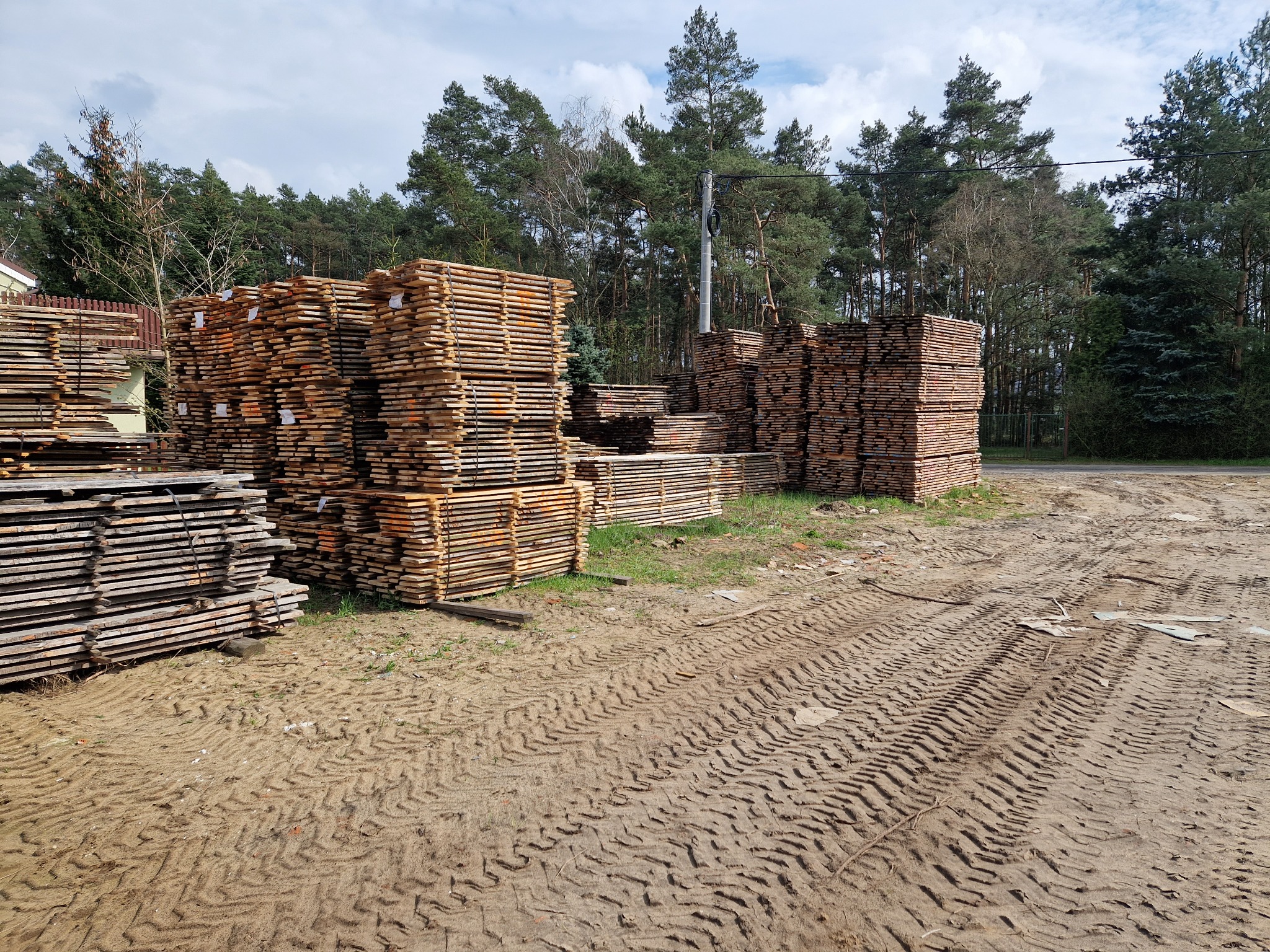 Stosy świeżo przetartego drewna ułożone na placu z widocznymi śladami opon, w tle las i fragment budynku.