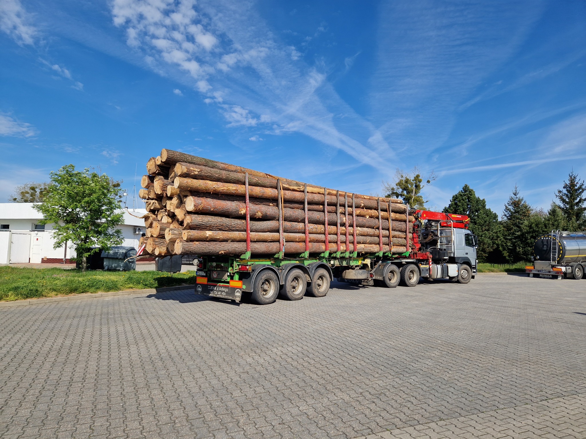 Ciężarówka z naczepą załadowana okrąglakami drewna na placu, widoczne zabezpieczenia ładunku, błękitne niebo w tle.