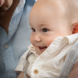 DUALART_PHOTOGRAPHY - Uśmiechnięty niemowlak w jasnej koszuli z drewnianymi guzikami i szarych szelkach, trzymany przez osobę w błękitnej koszuli.