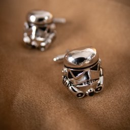 DUALART_PHOTOGRAPHY - Dwa srebrne spinki do mankietów w kształcie hełmów szturmowców z Gwiezdnych Wojen leżą na brązowym, teksturowanym tle.