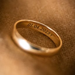 DUALART_PHOTOGRAPHY - Złota obrączka ślubna z wygrawerowaną datą 01.04.2023 na beżowym tle.