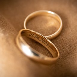 DUALART_PHOTOGRAPHY - Dwa złote pierścionki ślubne na beżowym, teksturowanym tle. Na wewnętrznej stronie jednego z pierścionków wygrawerowana data: 01.04.2023.