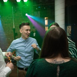 DUALART_PHOTOGRAPHY - Grupa młodych ludzi w koszulach tańczy w ciemnym pomieszczeniu oświetlonym przez zielone i niebieskie światła punktowe. W tle ceglana ściana i kolumna.